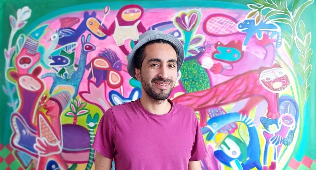 Artist Nima Javan wearing pink t-shirt and grey hat standing in front of one of his artworks with blue and pink creatures, trees and a blue and yellow border